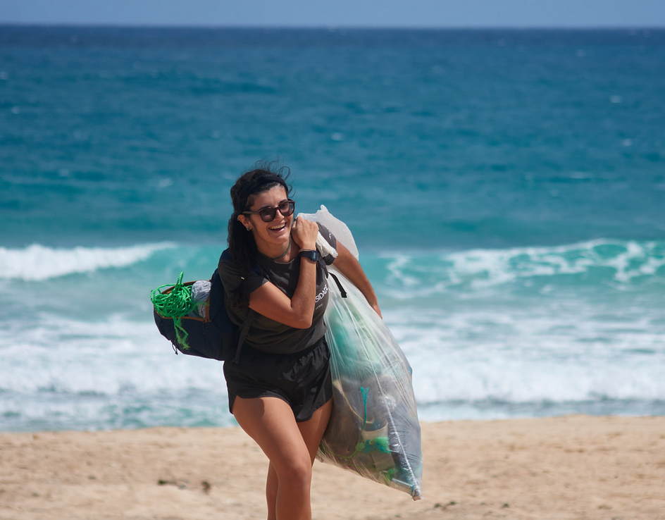 A smiling woman carries a bag of trash on a beach.